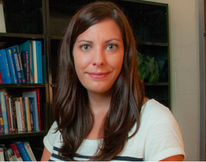 A smiling woman with long brown hair and light eyes wears a striped shirt, with bookshelves in the background.