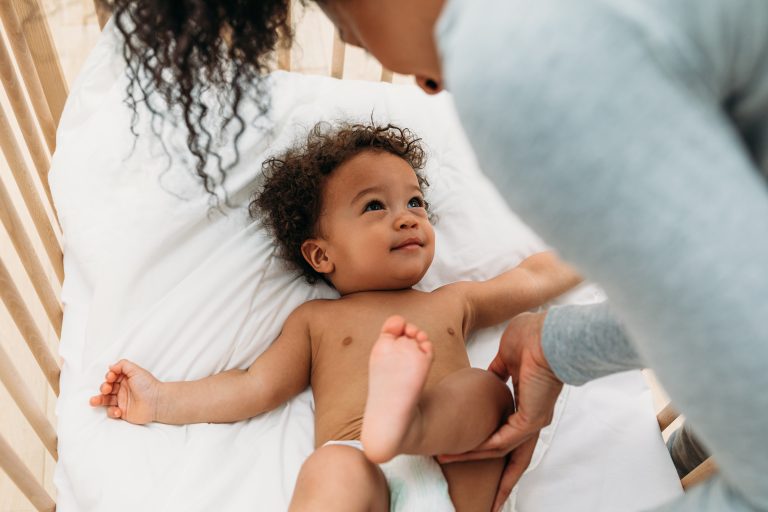 A parent gently holds their smiling baby's legs in a crib as the baby looks up during a diaper change.