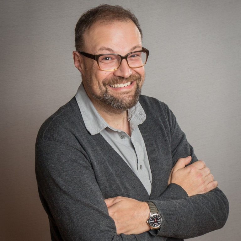 Professional headshot of a smiling man with a beard and glasses, wearing a grey shirt and dark grey cardigan.