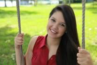 A smiling young woman with long dark hair sits on a swing outdoors.