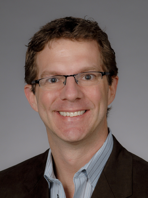 Smiling man with brown curly hair and glasses, wearing a striped blue shirt and brown jacket.