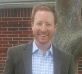 Professional headshot of a smiling man with reddish-brown hair wearing a blue checkered shirt and dark jacket.