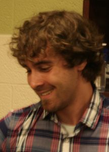 A smiling man with curly brown hair and a light beard wears a blue, red, and white plaid shirt.