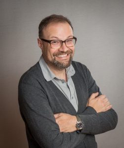 Professional portrait of a smiling man with a beard and glasses, wearing a grey cardigan and shirt, with his arms crossed.
